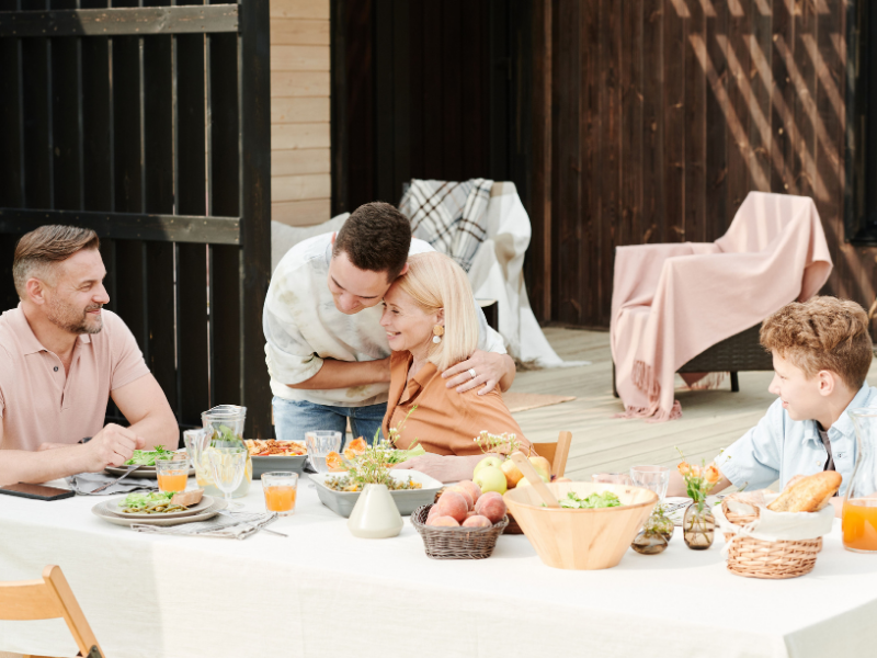 Family gathering outdoors with a table of food, including salads, bread, and drinks, in a backyard setting with sunlight and outdoor furniture.