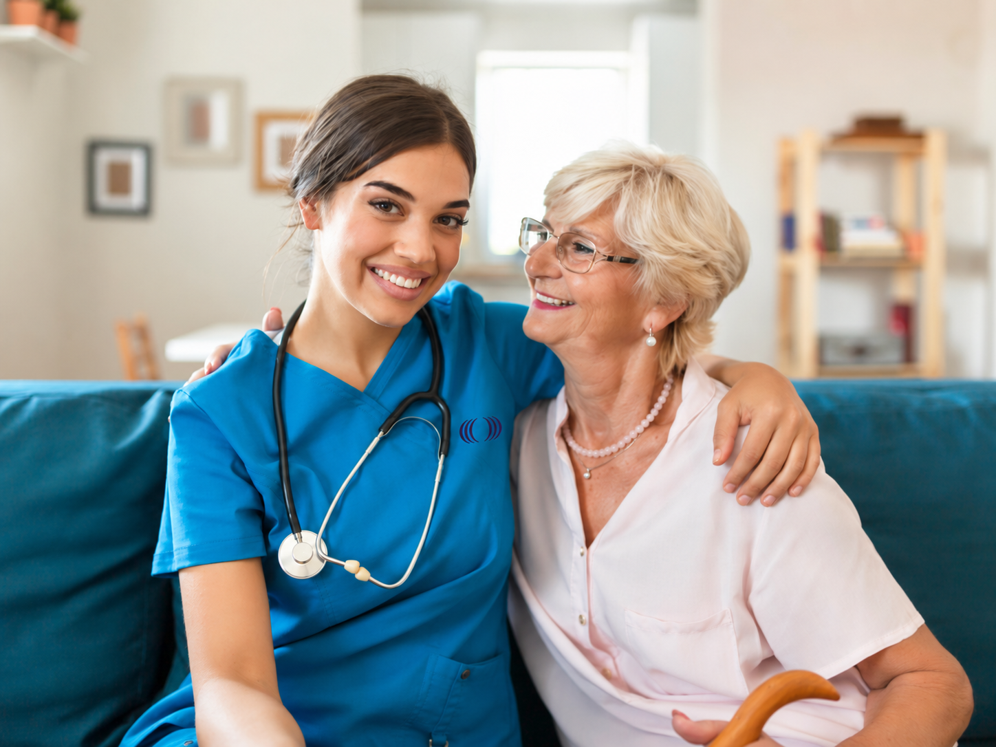 A young female healthcare worker wearing scrubs and a stethoscope hugging an elderly woman with glasses and white hair sitting on a couch.