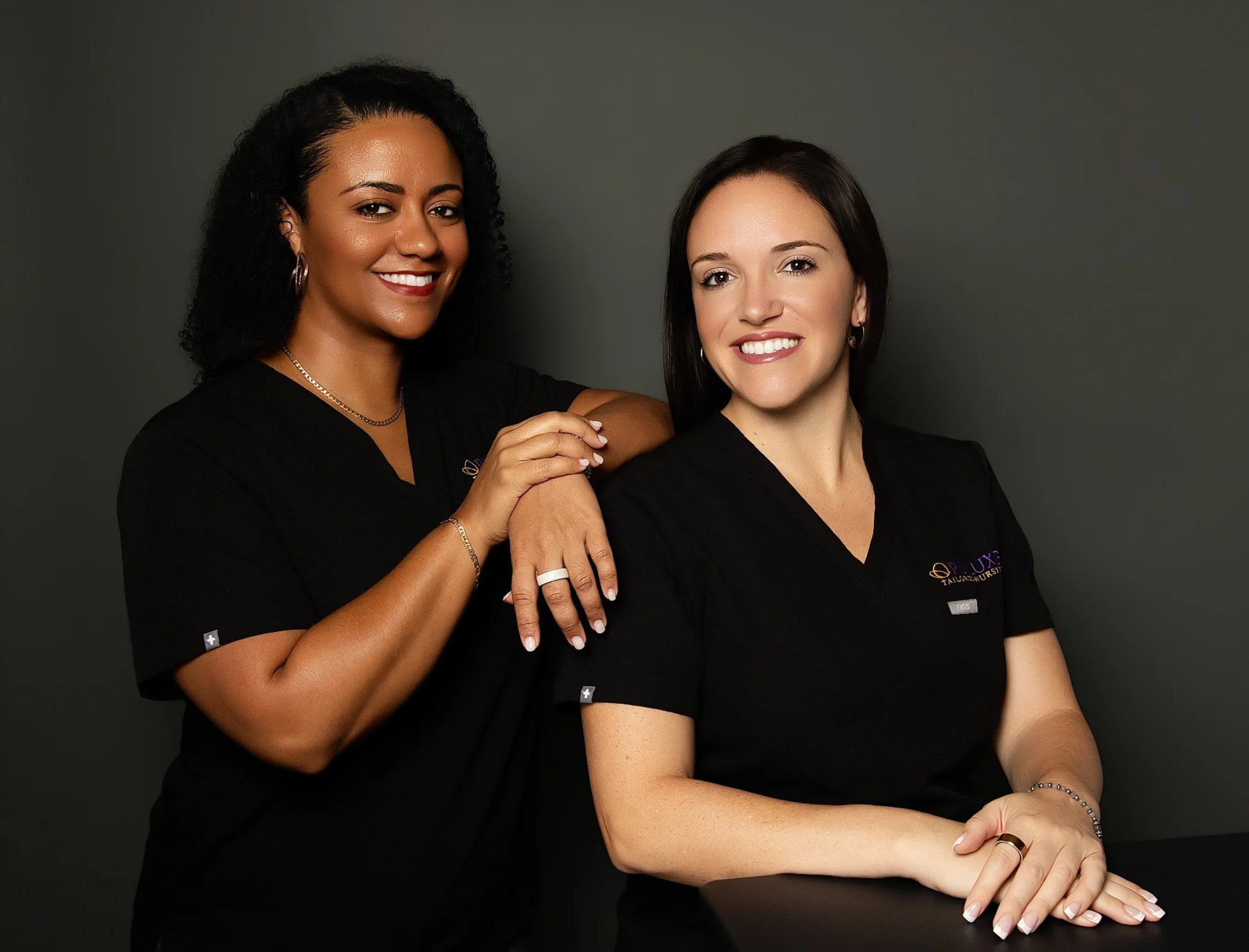Two smiling women wearing black shirts against a dark background, with one woman's arm resting on the other's shoulder.