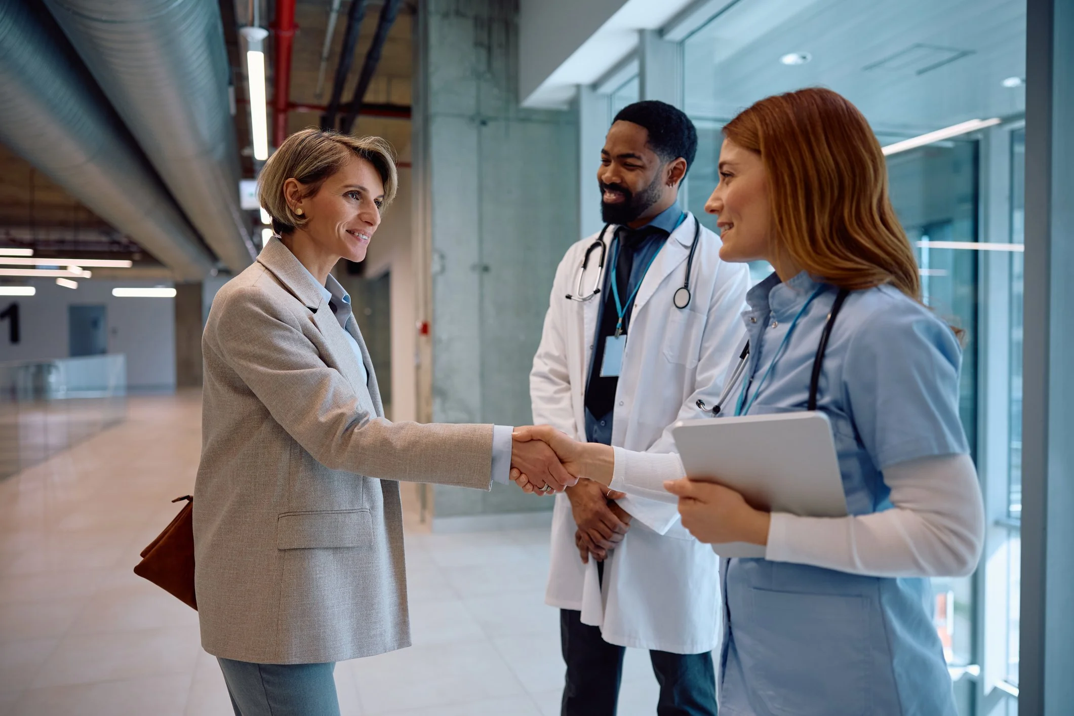 A woman in a beige blazer shaking hands with a female healthcare professional holding a tablet, while a male healthcare professional stands nearby in a modern hospital hallway with large windows.