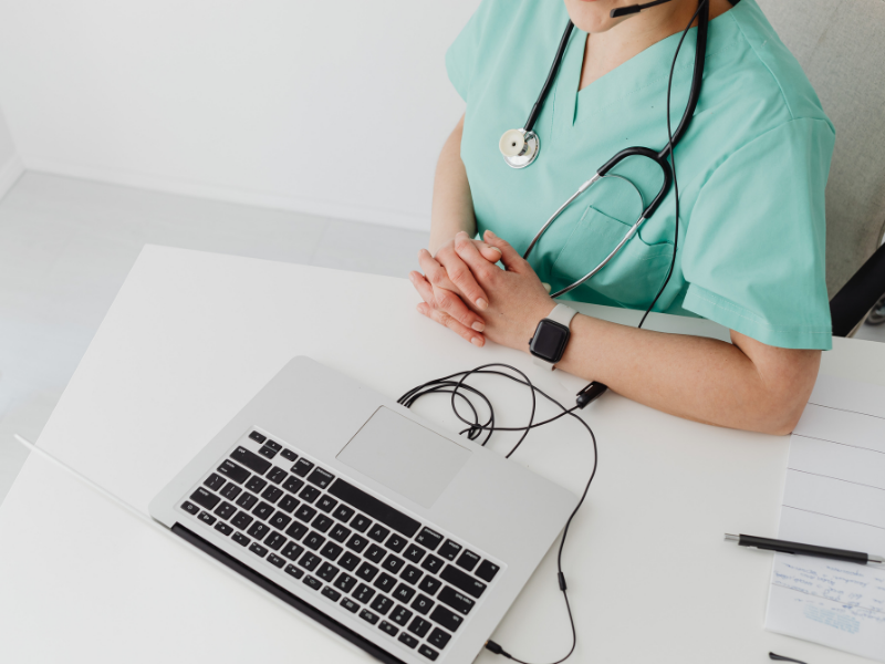 Healthcare professional in scrubs sitting at a desk with a laptop, wearing a headset and a smartwatch, with a notepad and pen nearby.