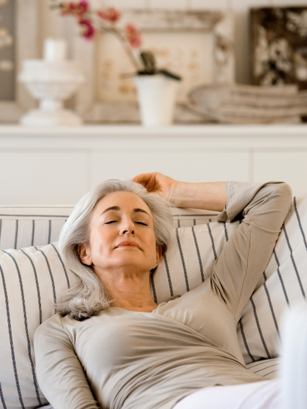 An elderly woman with gray hair relaxing on a striped couch with her eyes closed and one arm behind her head in a cozy living room.