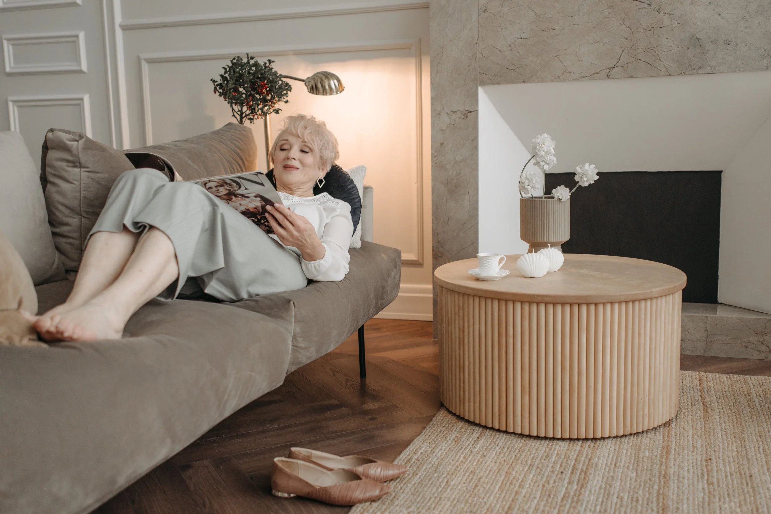 An elderly woman with blonde hair lies on a gray couch reading a magazine. She appears relaxed and content. The room has neutral-colored decor with a marble accent wall, a wooden side table with white vases and flowers, and a coffee cup. Shoes are placed on a round rug on a dark wooden floor. A small Christmas tree with lights and ornaments is visible in the background.