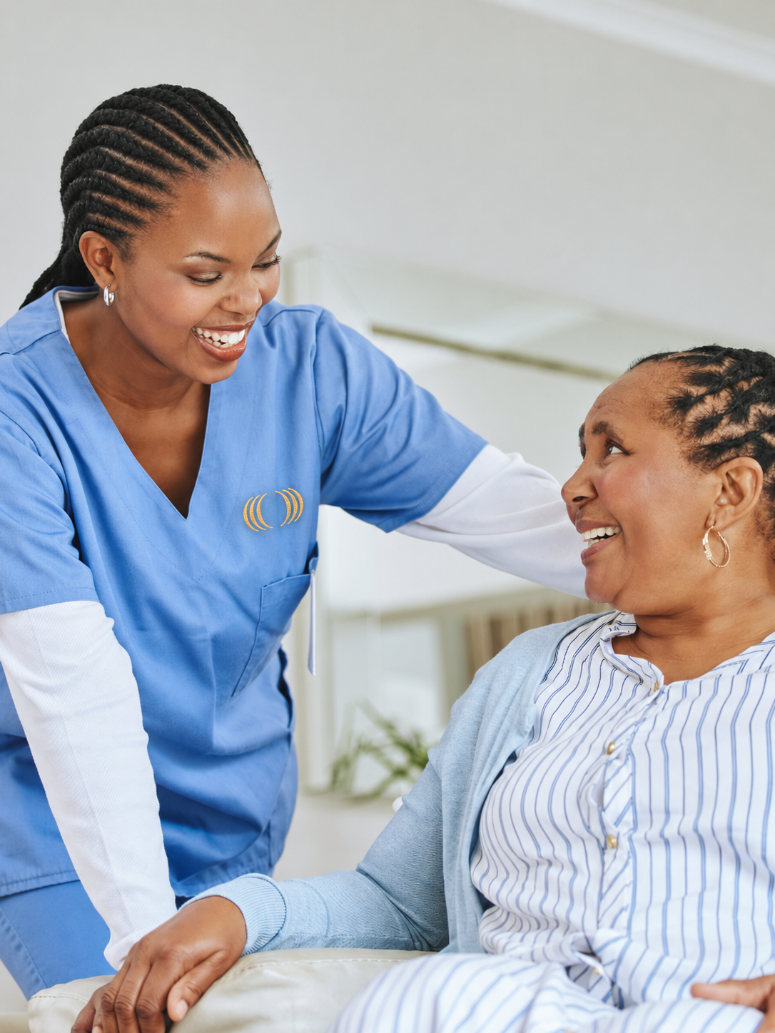 A healthcare professional, wearing blue scrubs, smiling and engaging with a patient, a woman sitting in a chair, in a medical setting.