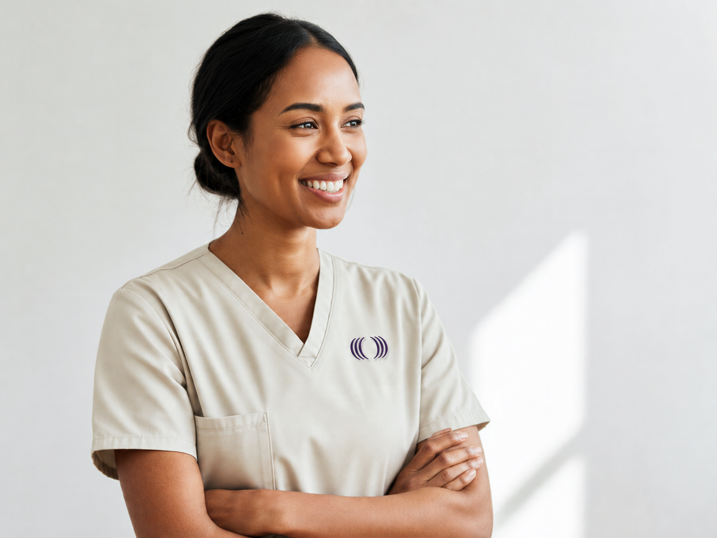 A smiling woman in beige medical scrubs with a purple logo on the chest, standing with her arms crossed against a white background.