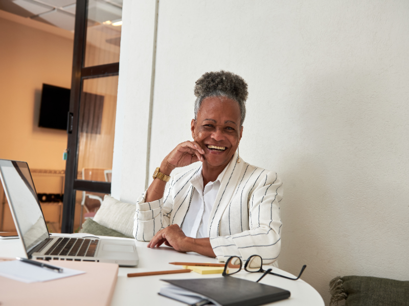 Smiling woman with short gray hair wearing a white blazer with black stripes, sitting at a desk with a laptop, eyeglasses, notebook, and pens in an office or workspace.