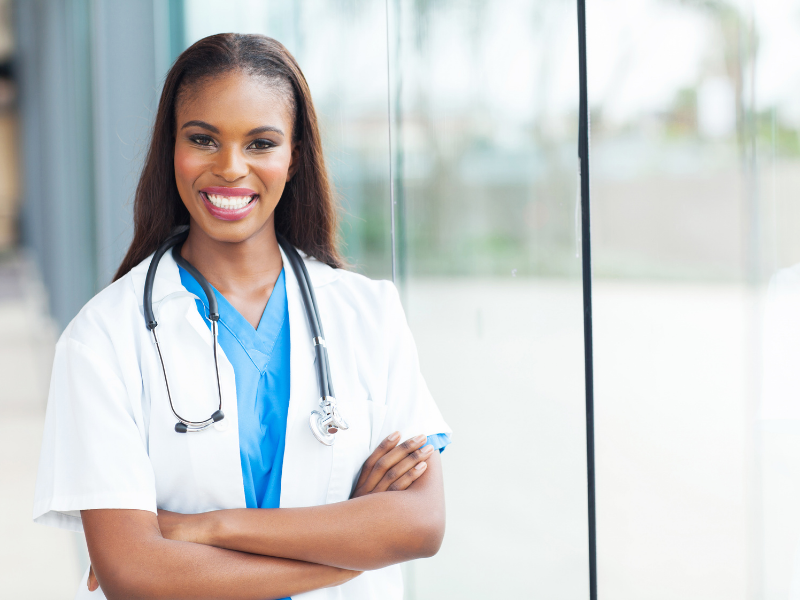 Smiling female healthcare professional wearing a white lab coat and blue scrubs, with a stethoscope around her neck, standing with arms crossed in front of a glass window.