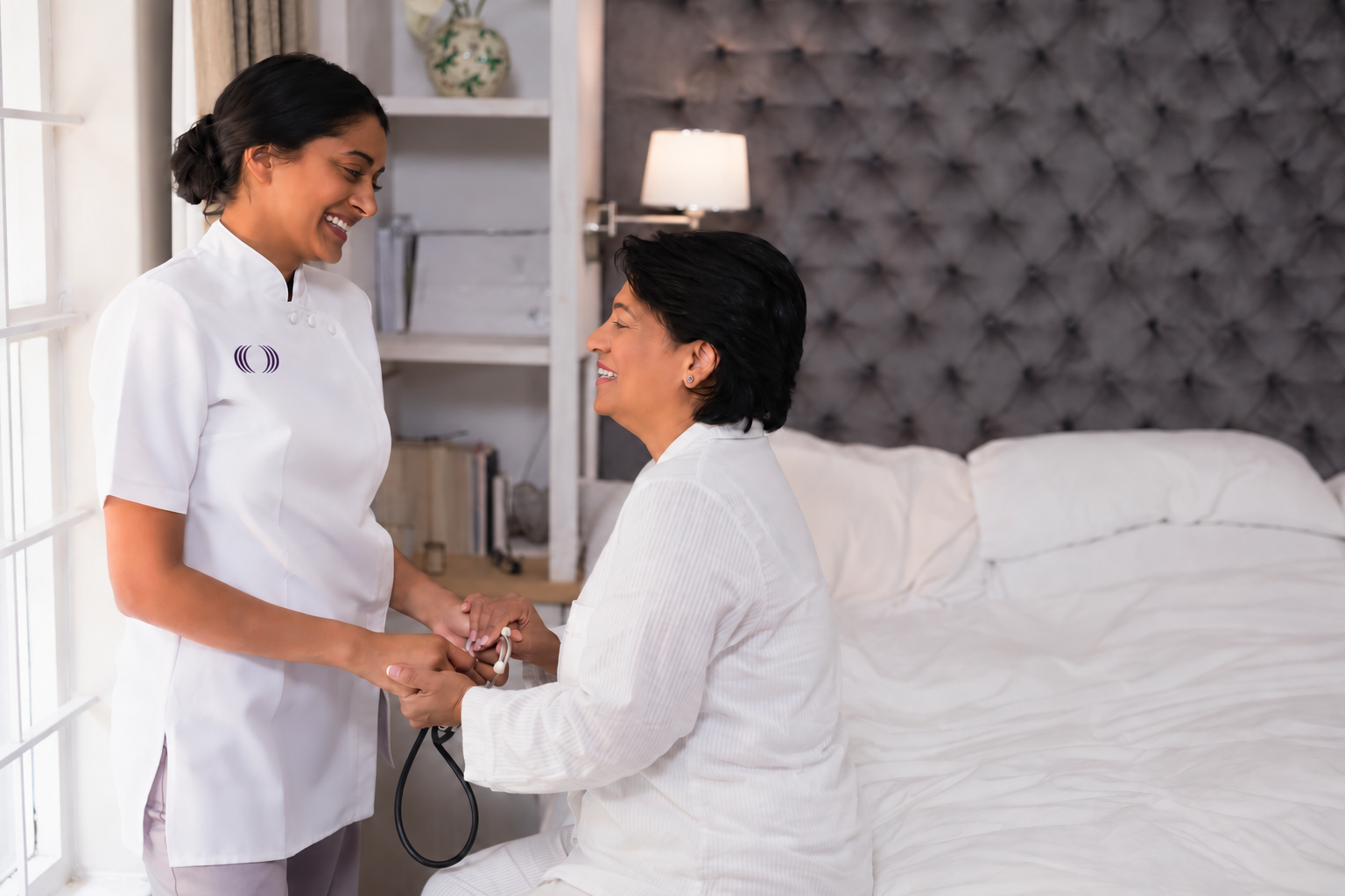 A healthcare professional and a patient smiling and holding hands in a bright, cozy bedroom.