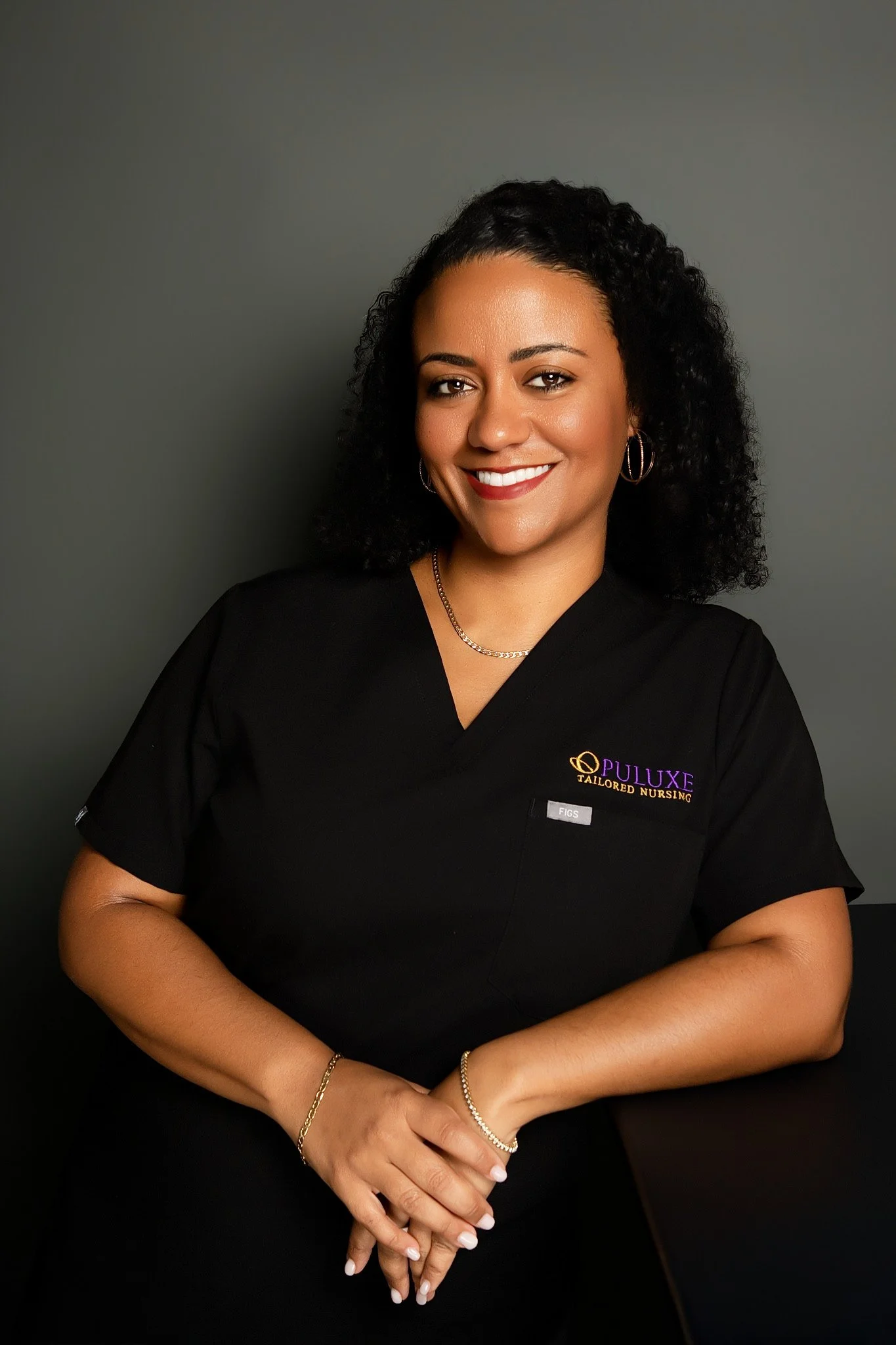 A woman smiling in a black medical uniform with gold jewelry, standing against a gray background.