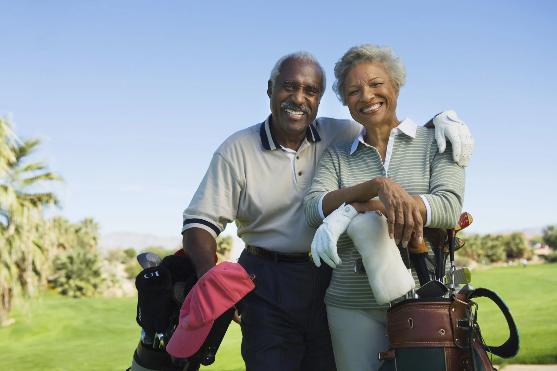 Two smiling seniors, a man and a woman, standing outdoors on a golf course with golf clubs and bags, enjoying a sunny day.