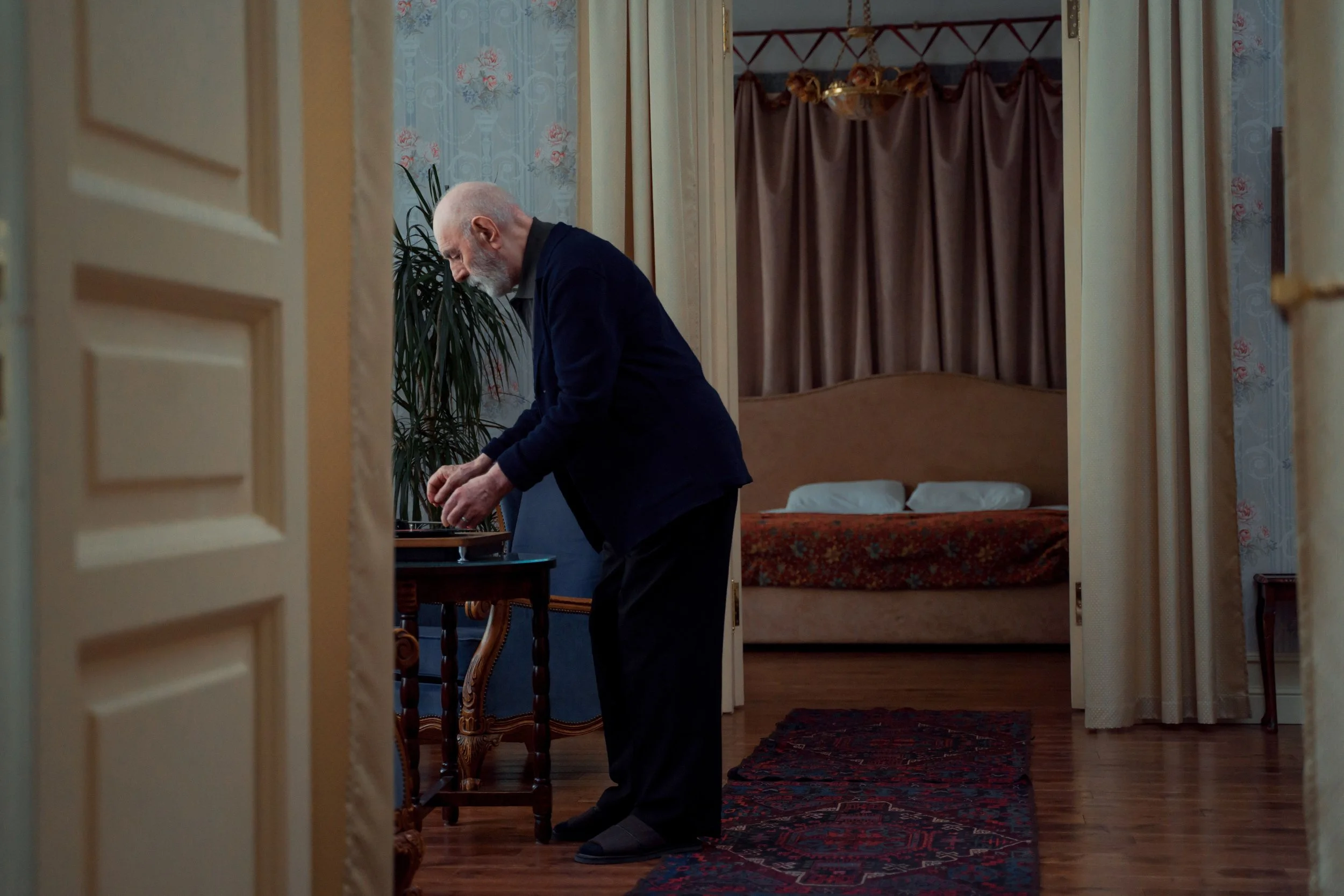 A bald man with a beard wearing a navy suit leans over and writes at a small table in a room with floral wallpaper, a bed with a floral blanket, and a large curtain in the background.