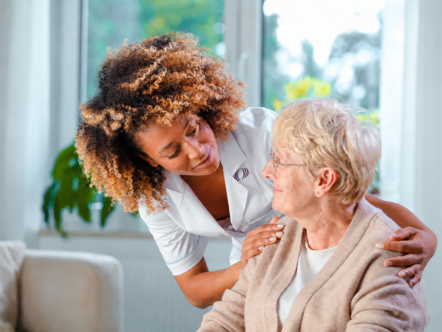 A healthcare professional comforting an elderly woman in a bright room with large windows and green plants in the background.