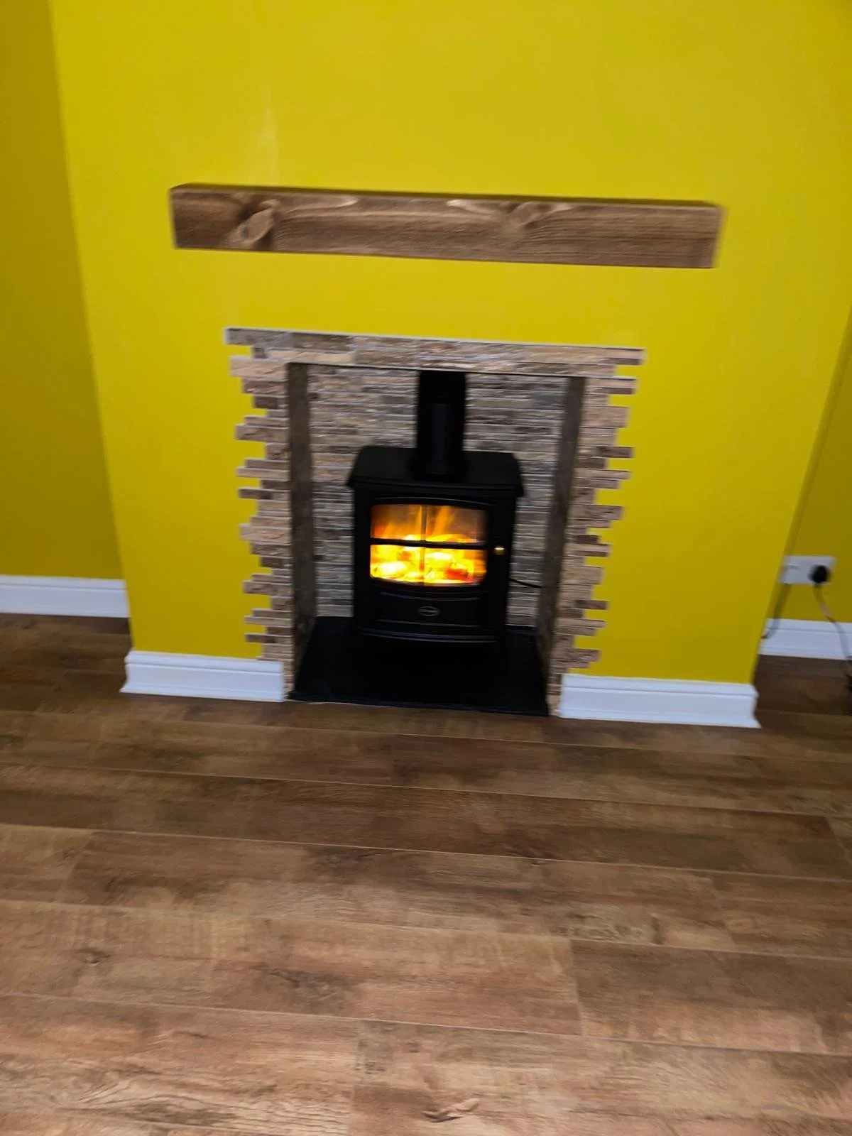Yellow wall with a small black wood stove inside a stone-lined fireplace opening and a wooden mantel above, with hardwood flooring.