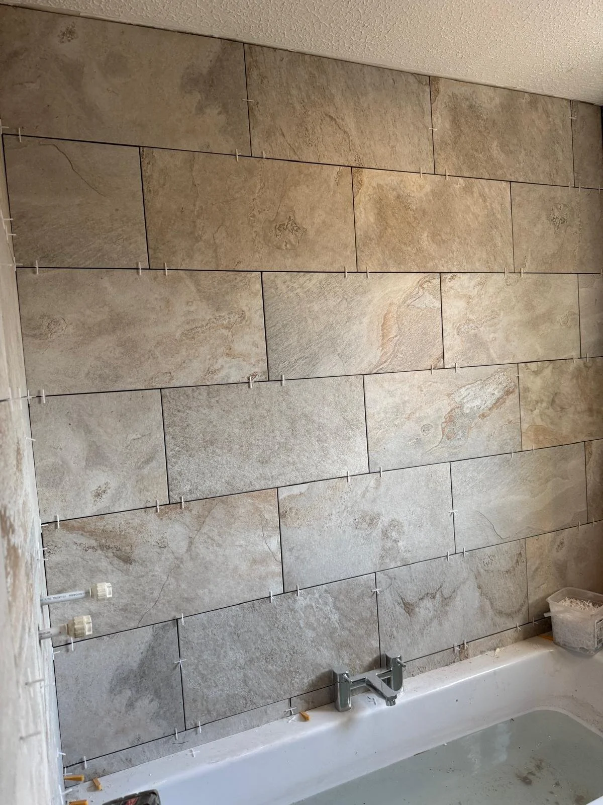 Bathroom wall with large beige and gray tiles being installed, with spacers in place, a bathtub at the bottom, and plumbing fixtures visible.
