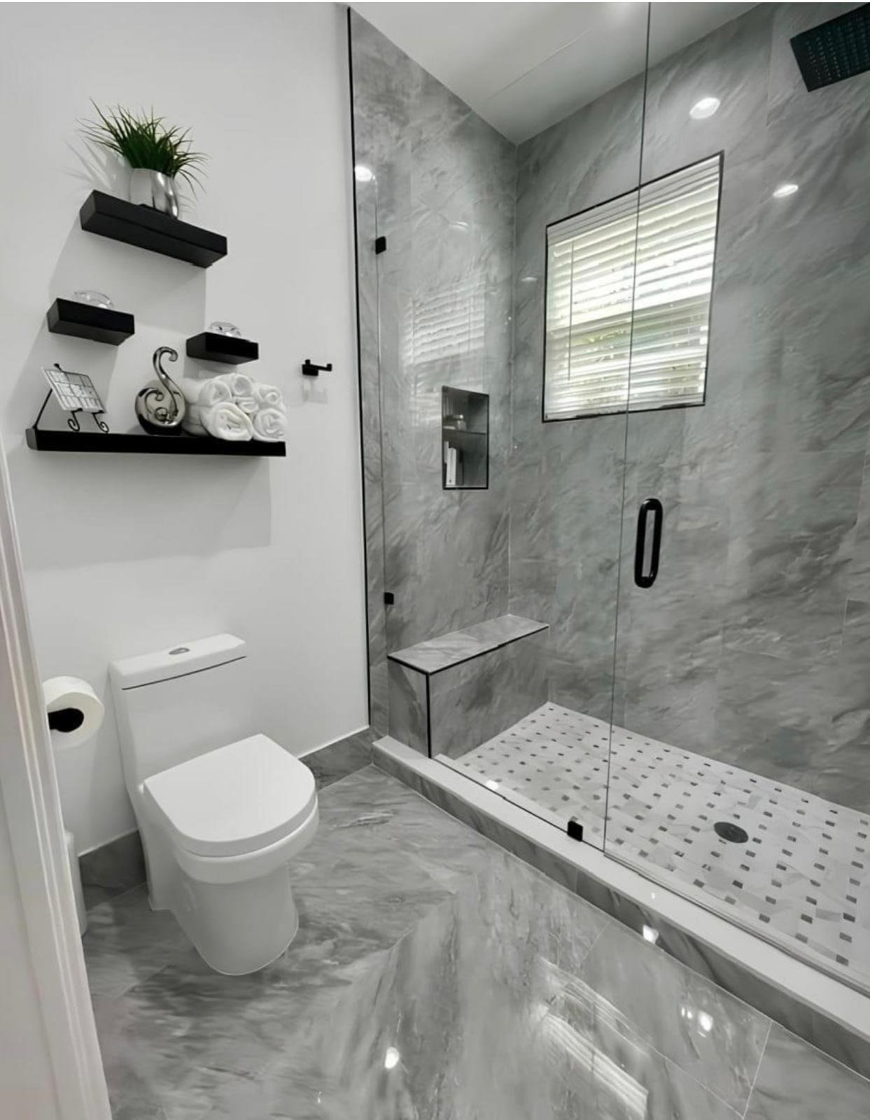 Modern bathroom with a white toilet, black wall-mounted shelves with decorative items, and a large glass-enclosed walk-in shower with marble walls and a window.