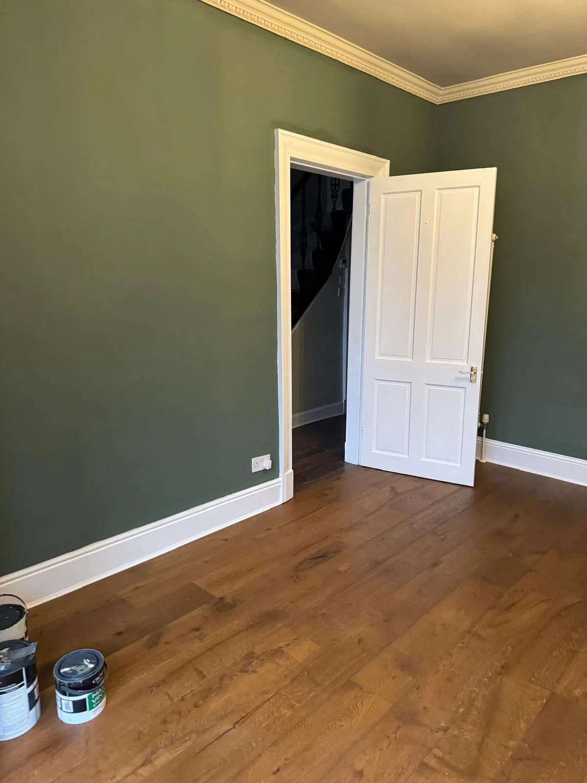 A room with green walls, a white trim, and a wooden floor. An open white door leads to a staircase area. There are cans of paint on the floor in the corner.