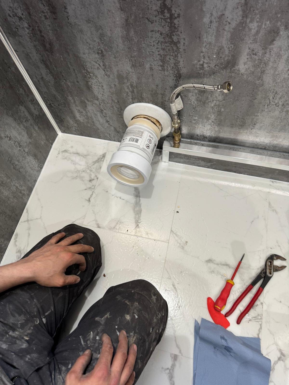 A person kneeling on a white marble-patterned floor, working on a plumbing connection near the wall. The person's hands are resting on their knees. There are tools including pliers and a screwdriver on the floor, and a section of PVC pipe and plumbing fittings are visible near the wall.