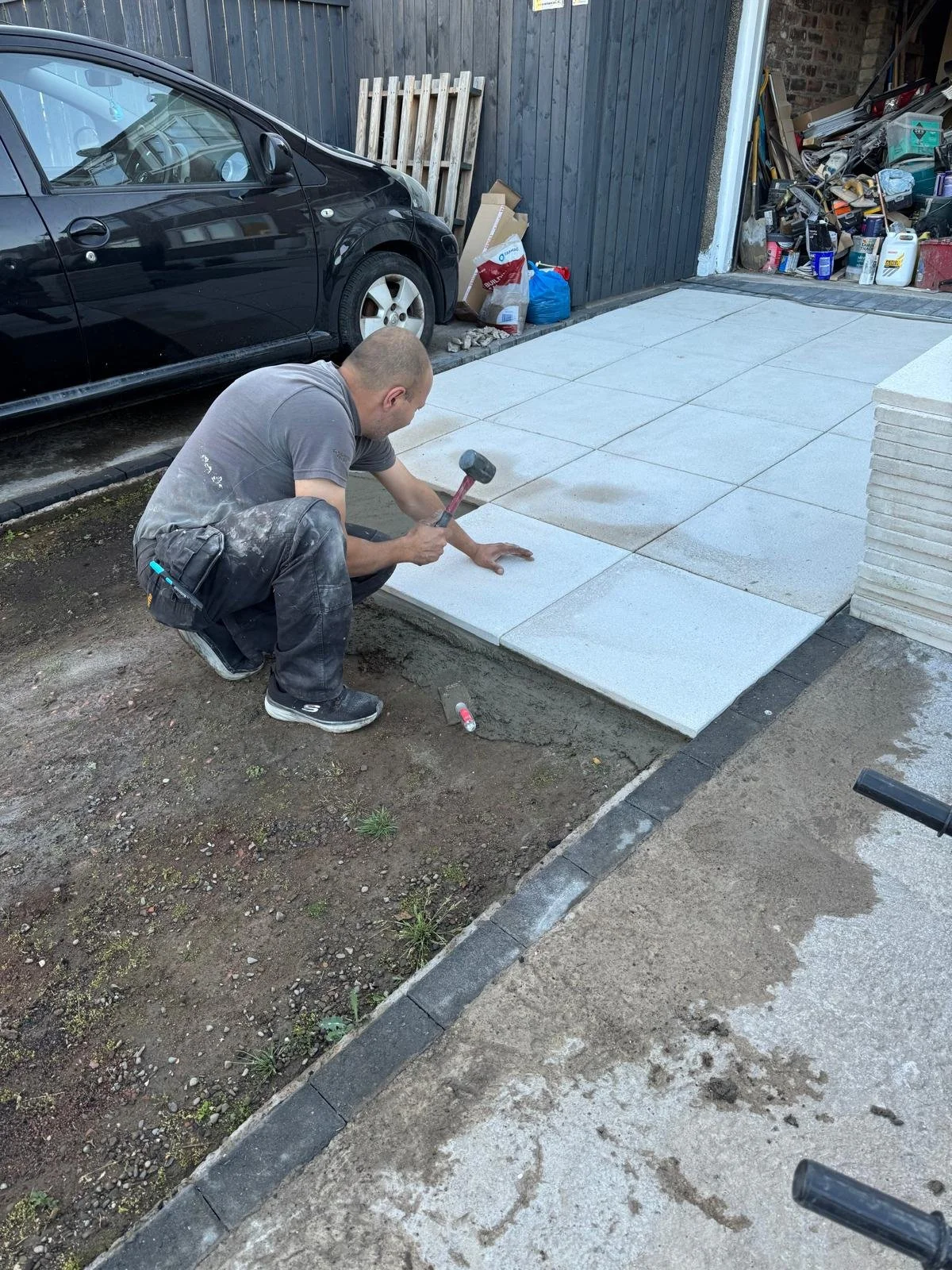 A man installing light gray concrete paving slabs on a walkway in a residential yard. The man is squatting and holding a hammer, with various tools and materials nearby. There is a black car parked next to the yard, and a wooden fence with a pile of bags and boxes in the background.
