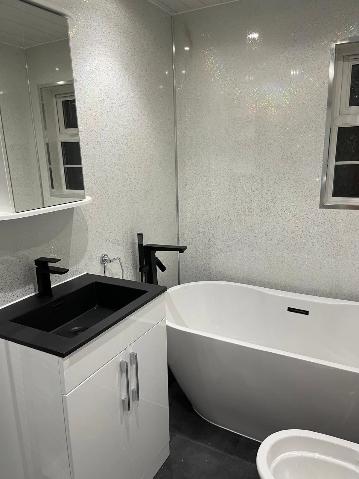 A modern bathroom with a white bathtub, black sink, and white cabinet; black faucet fixtures, glittery wall and window reflections.