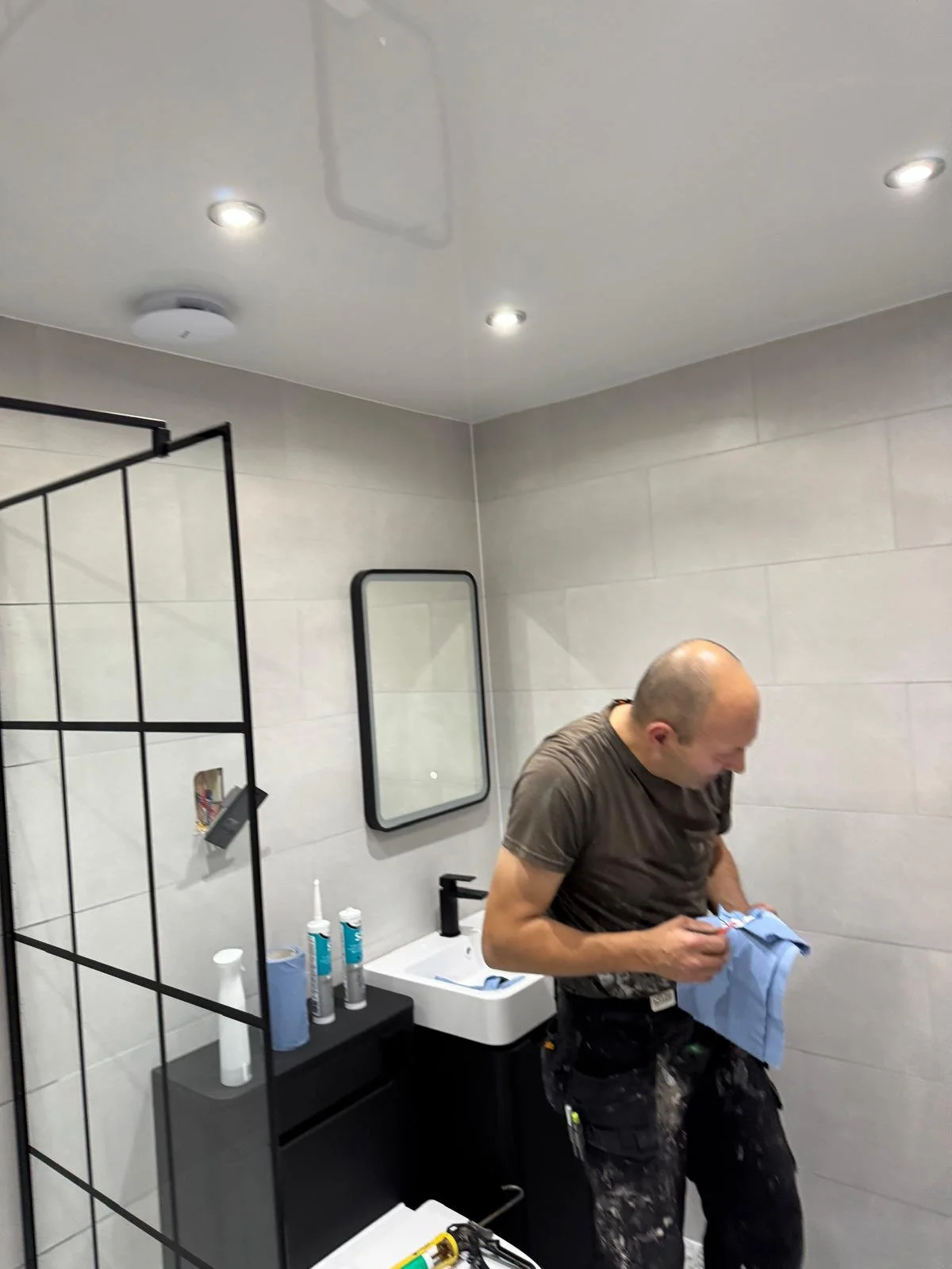 A man in a bathroom, wearing a brown shirt and black work pants, holding a blue cloth, with various tools and cleaning supplies on the sink counter.