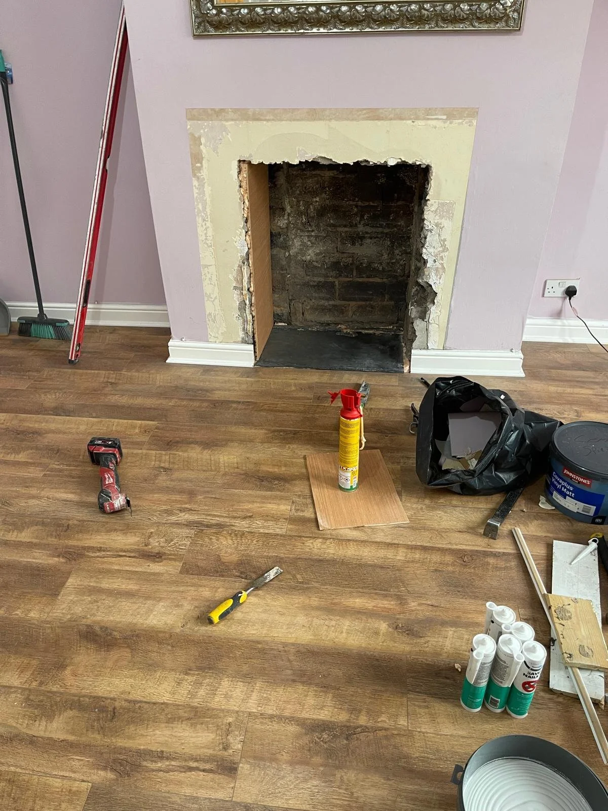 Room under renovation with removed fireplace, tools, and supplies on wooden floor, including a cordless drill, screwdriver, paint spray, and pipes.