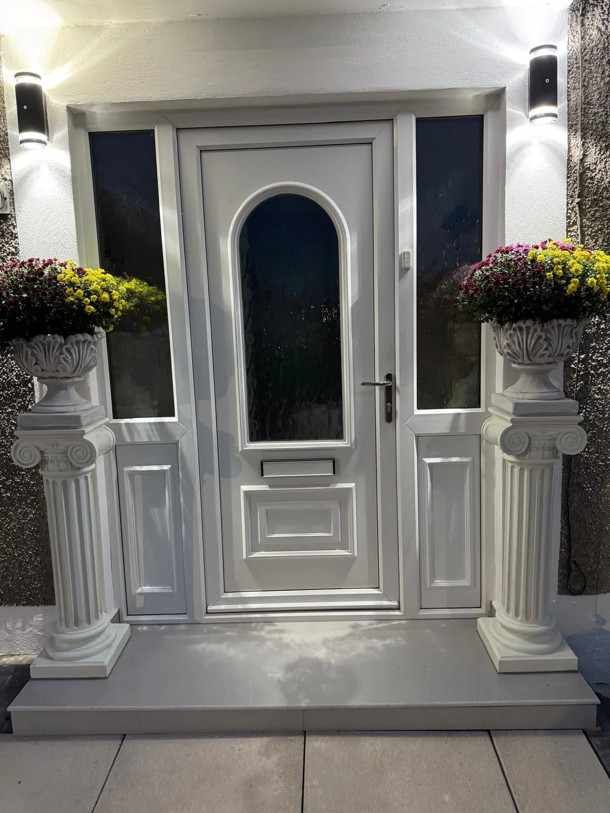A white door with frosted glass panels is flanked by two classical white pedestals with floral arrangements. The door is equipped with a handle and mail slot, and is illuminated by two modern exterior wall lights.