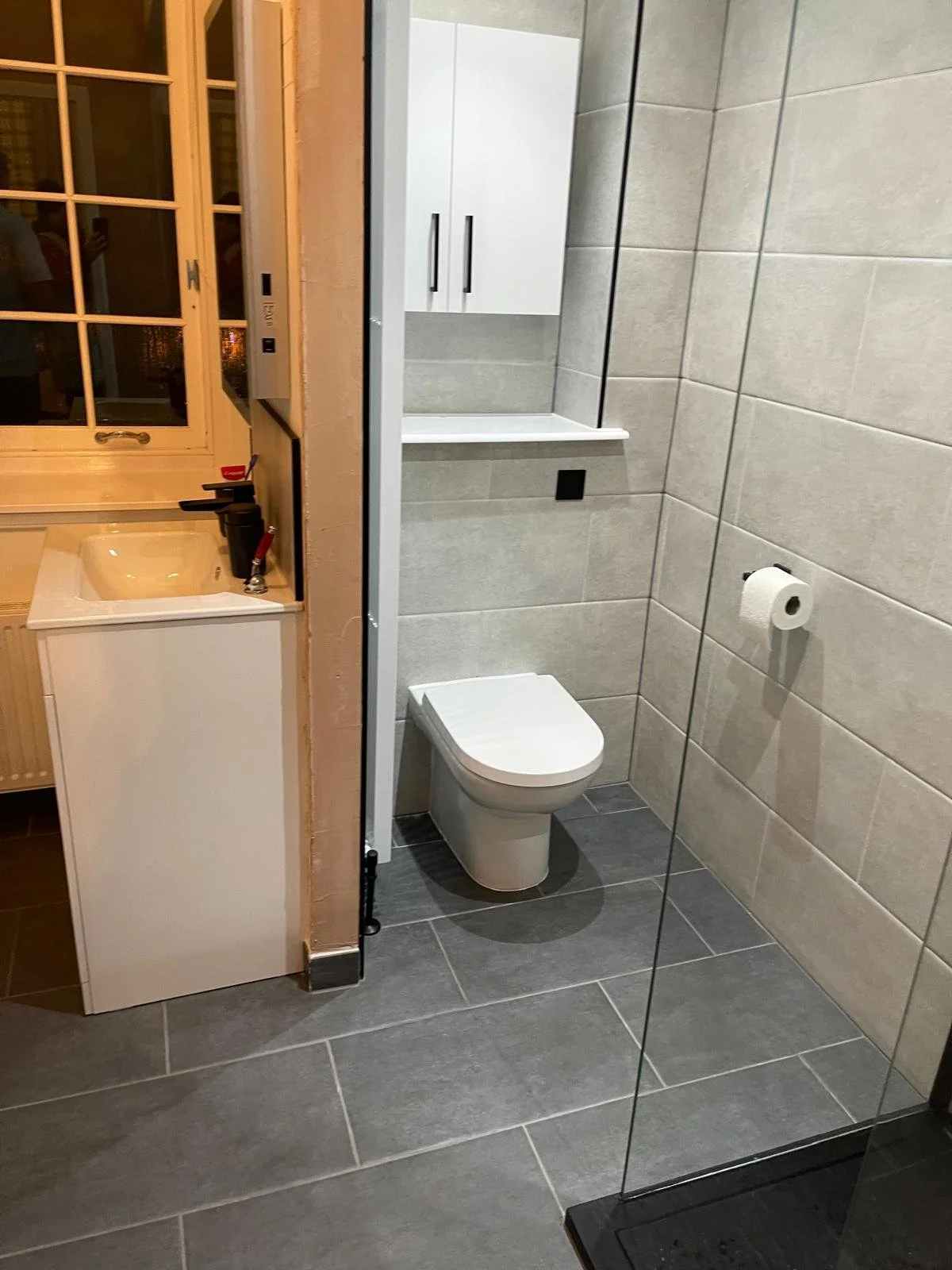 Bathroom with gray tiled walls and floor, a white toilet, a white wall-mounted cabinet, and a toilet paper roll on the wall.