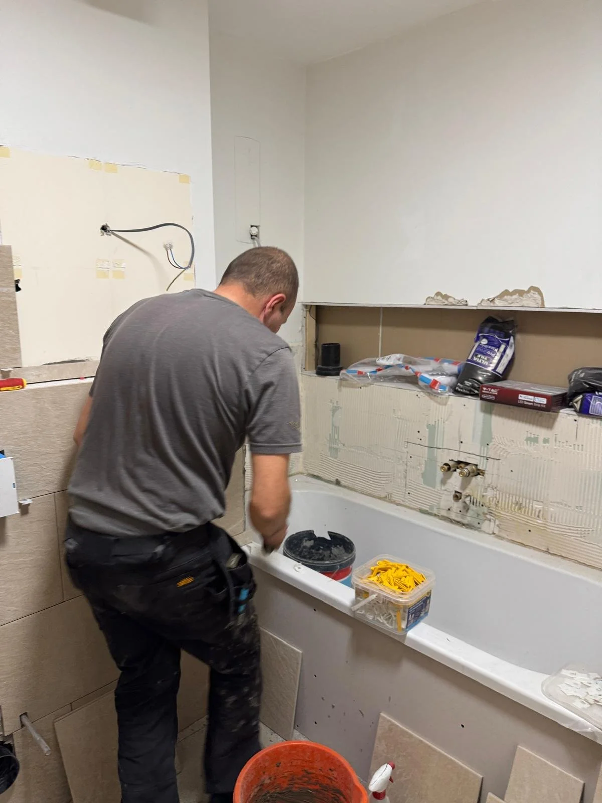 A man in a gray shirt and black pants working in a bathroom under renovation, installing or repairing the bathtub. There are construction materials, tools, and supplies around him, including a yellow hose and various boxes.