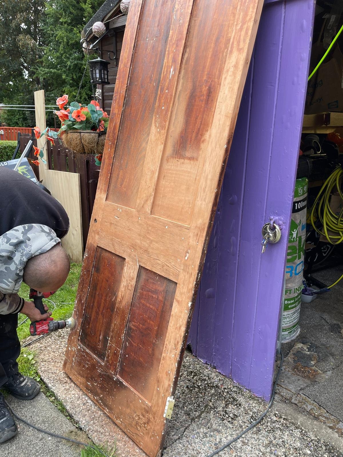 A person working with a power drill on a large, old wooden door propped up against a purple wall outside. The door has a keyhole and worn paint. Behind them, there is a background of trees, a fence with pink flowers, and a lantern hanging on the wall near the roof.