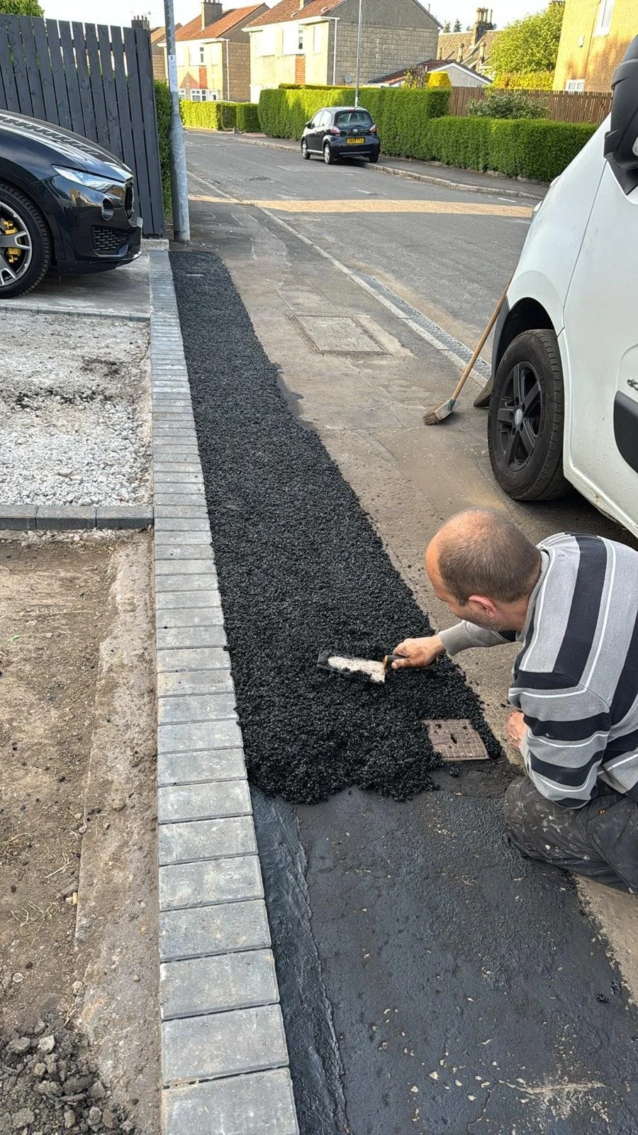 A man is laying down black asphalt with a trowel along the edge of a sidewalk, working on a new patch of pavement. A white van is parked nearby, and other cars and houses are visible in the background.