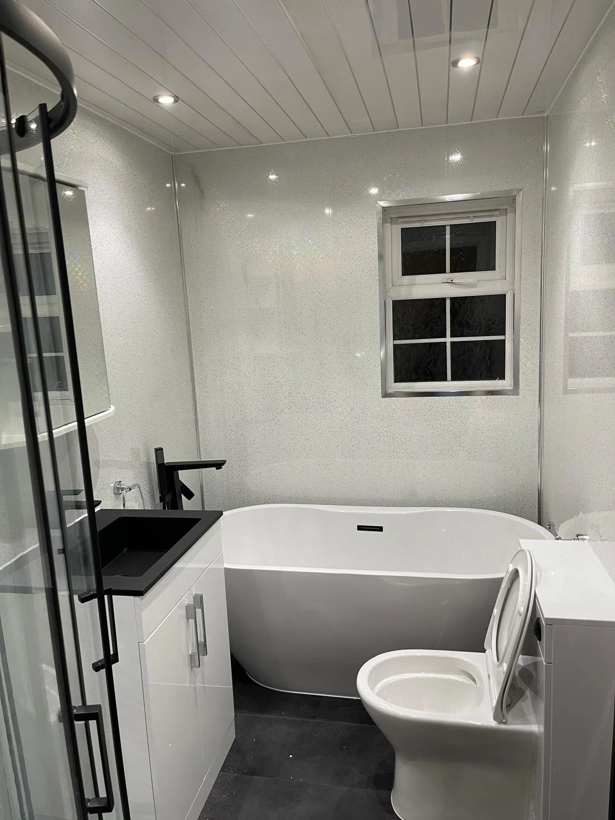 Modern bathroom with white walls, a black sink, a black faucet, a white bathtub, a toilet with the lid open, a small window, overhead lighting, and a washing machine.