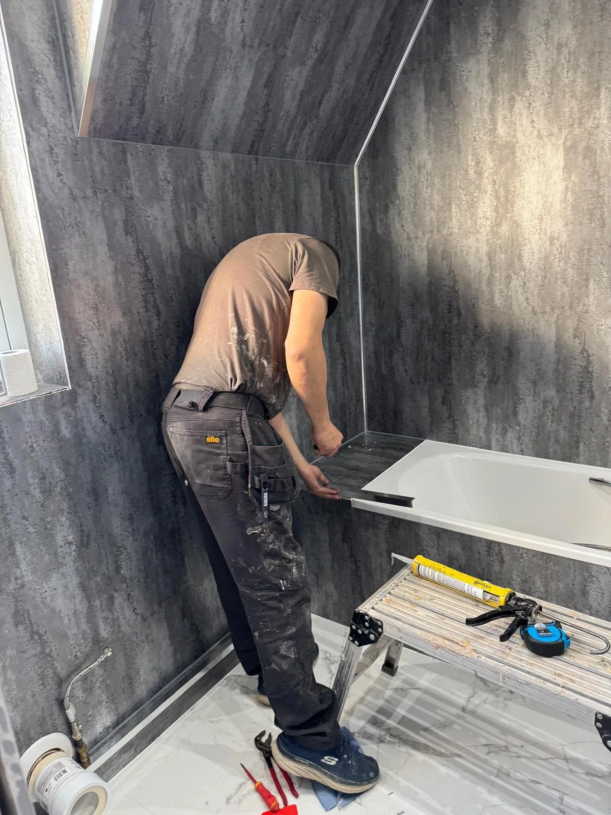 A worker installing or repairing a bathtub in a bathroom with dark gray textured walls. The worker is bending over, applying a sealant or adhesive to the edge of the bathtub. Tools and supplies like a caulking gun, measuring tape, and screwdrivers are on a small ladder nearby.