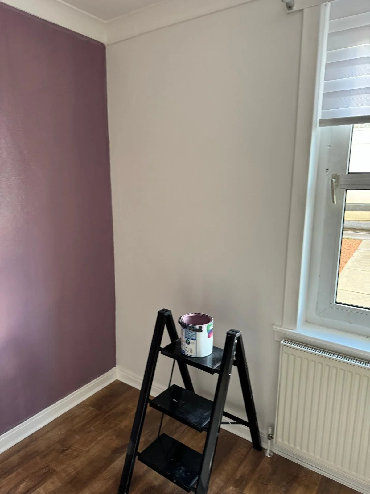 Interior of a room with two-tone wall paint, purple on the left and white on the right. A window with a partially open roller blind and a radiator below. A black stepladder with a paint can on the top step.