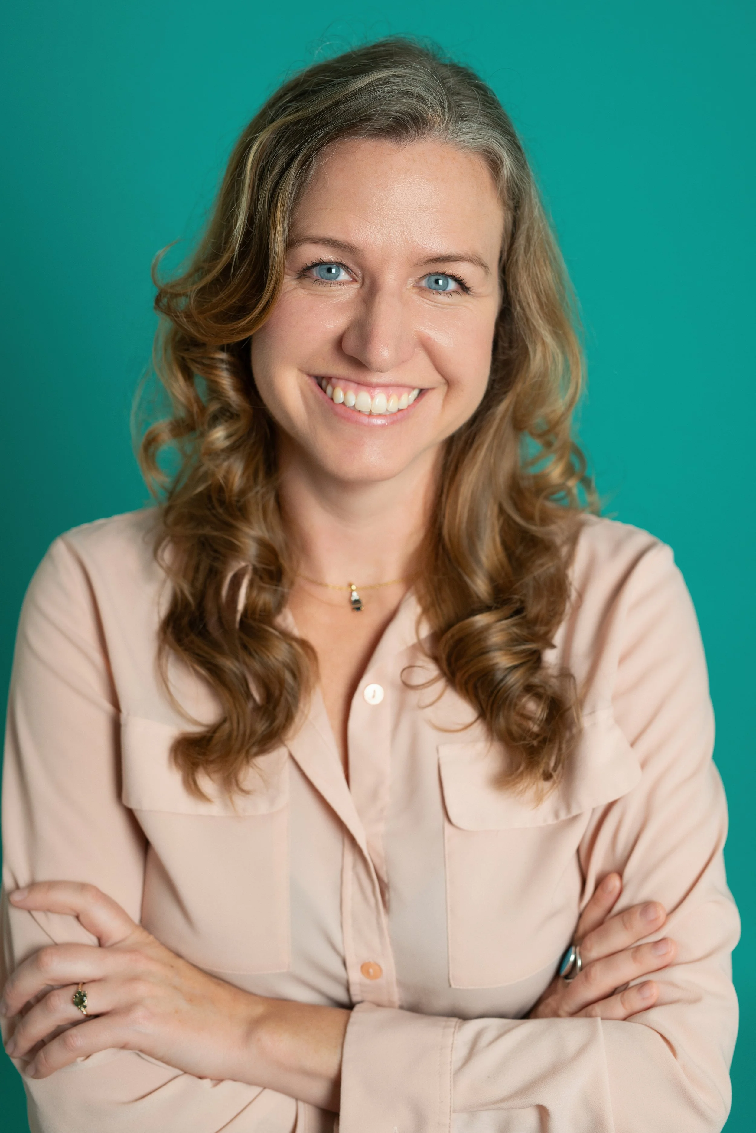 A smiling woman with light brown, curly hair wearing a light pink button-up shirt and a necklace, standing against a teal background.