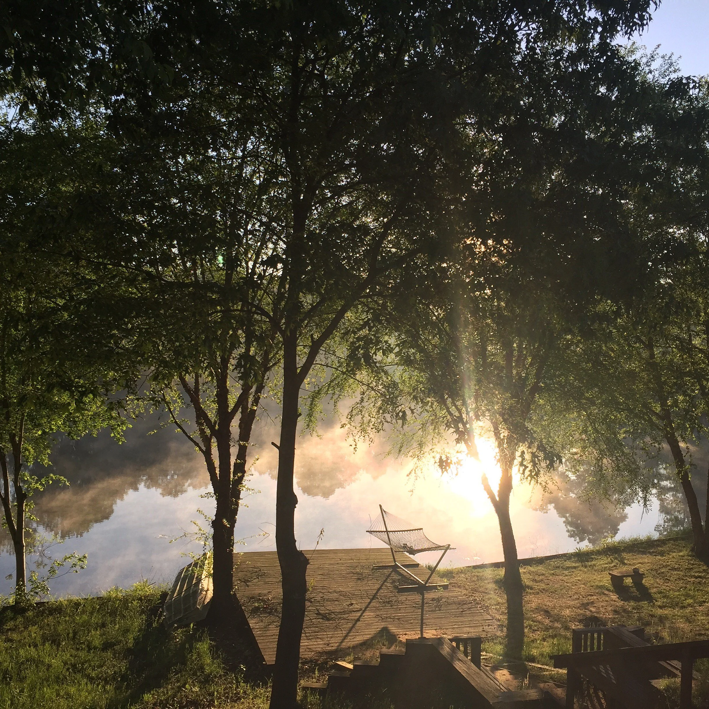 Sunset or sunrise through trees near a wooden deck with outdoor furniture.