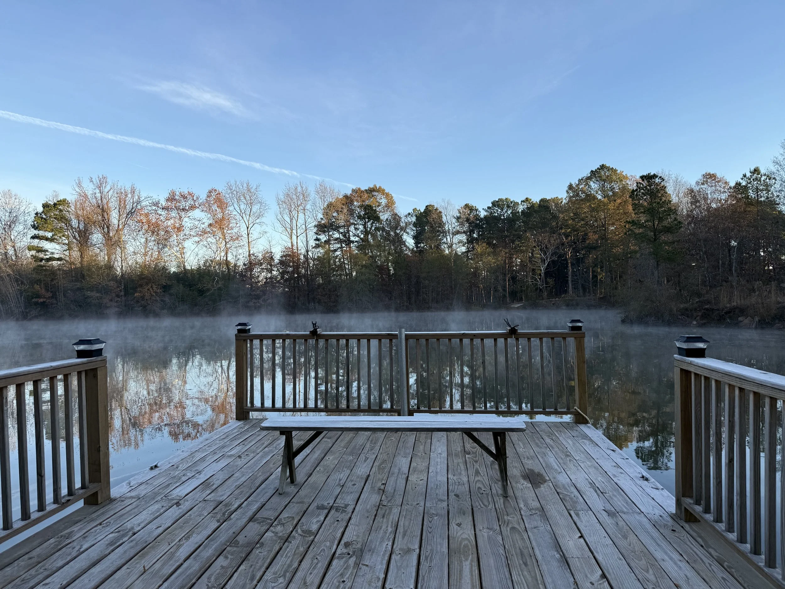 View of a misty lake from a wooden deck with benches, surrounded by trees with autumn foliage under a clear blue sky.