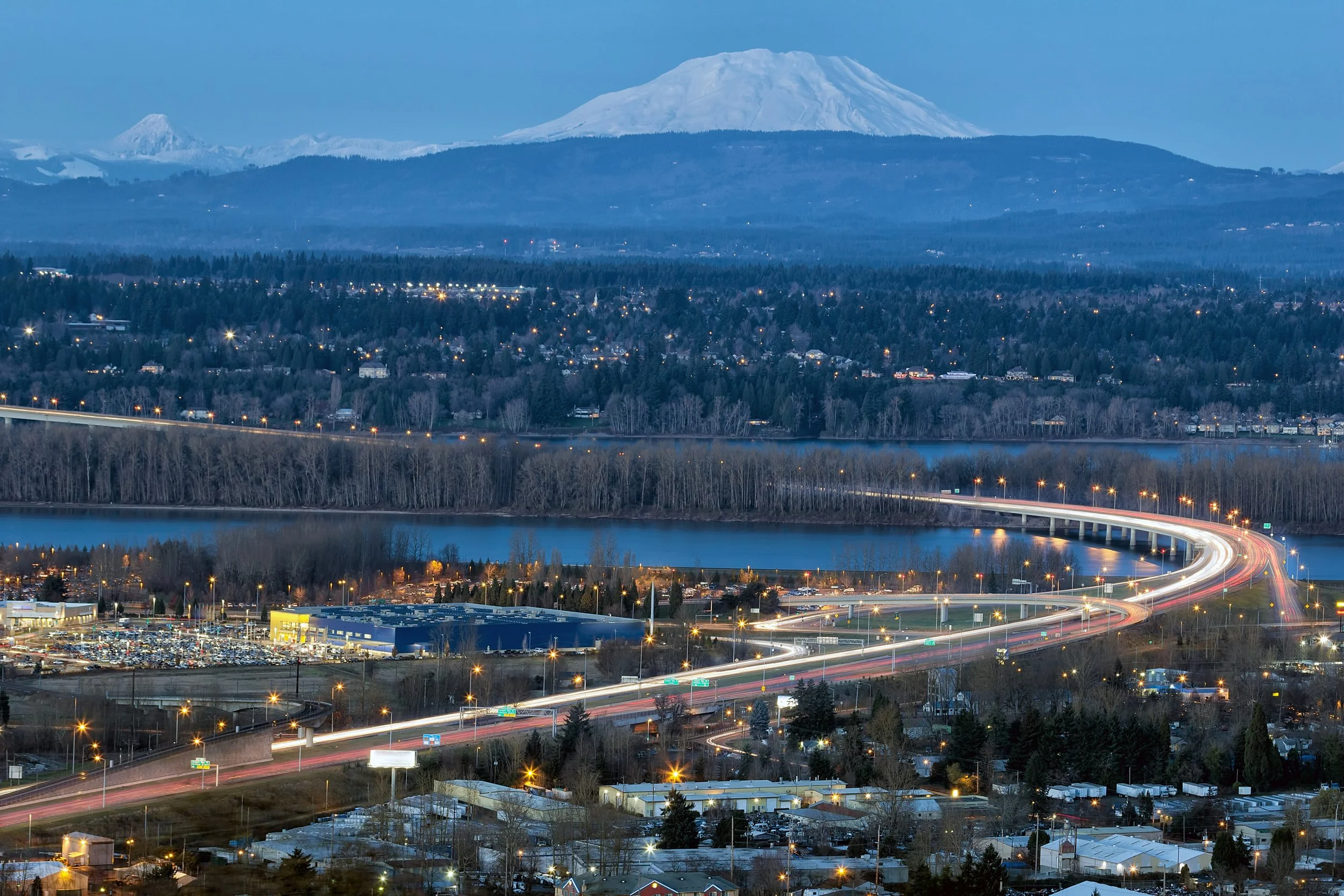 Nighttime cityscape with a large body of water in the foreground, a bridge with light trails from moving vehicles, a forested area, and snow-capped mountains, including Mount Hood in the background.
