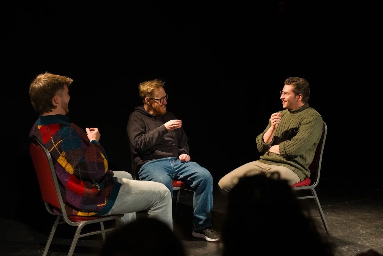 Three men sitting in chairs on a dark stage, engaged in a conversation or discussion, smiling and gesturing.