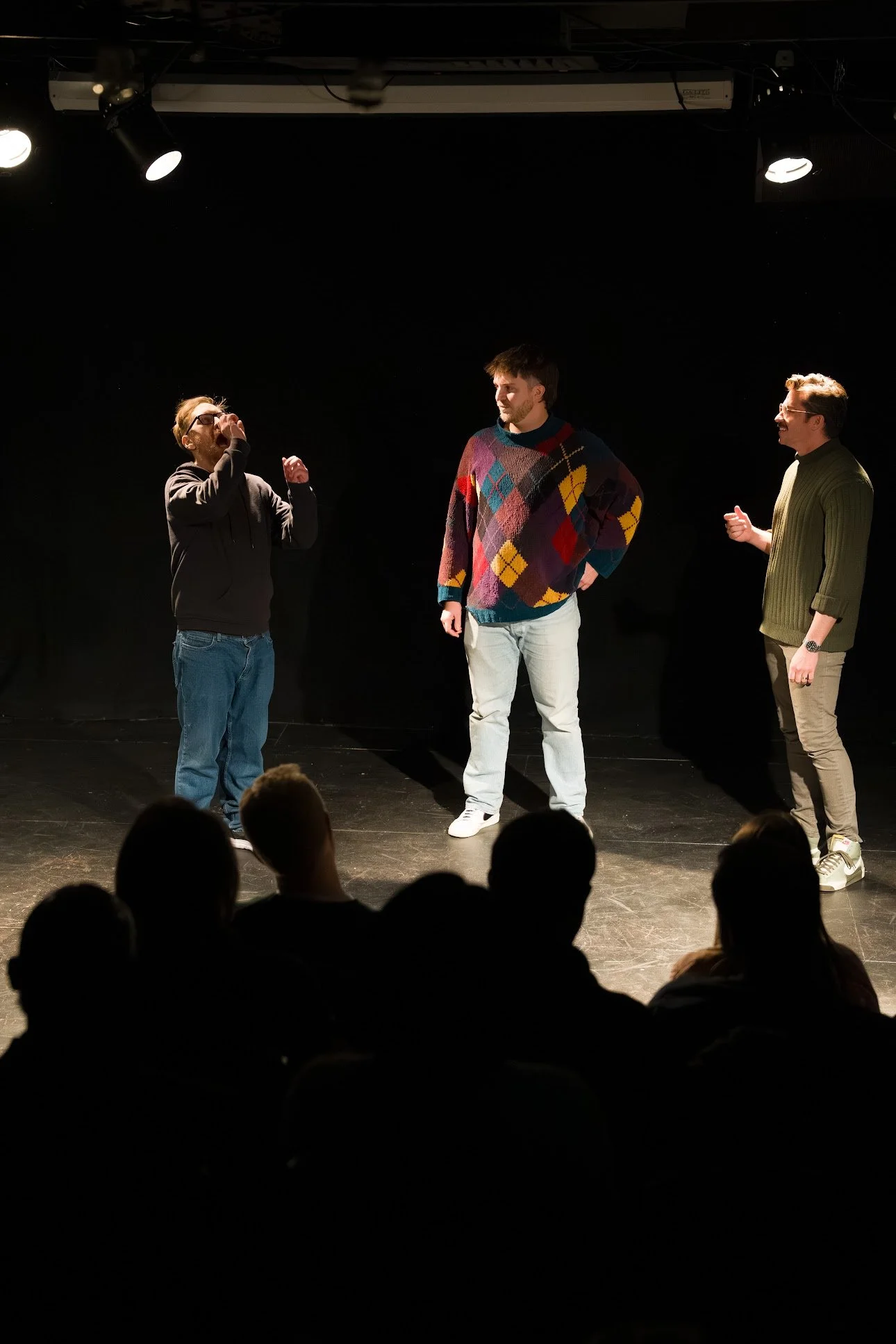 Three men performing a comedy routine or play on a dimly lit stage, with audience members visible in the foreground.