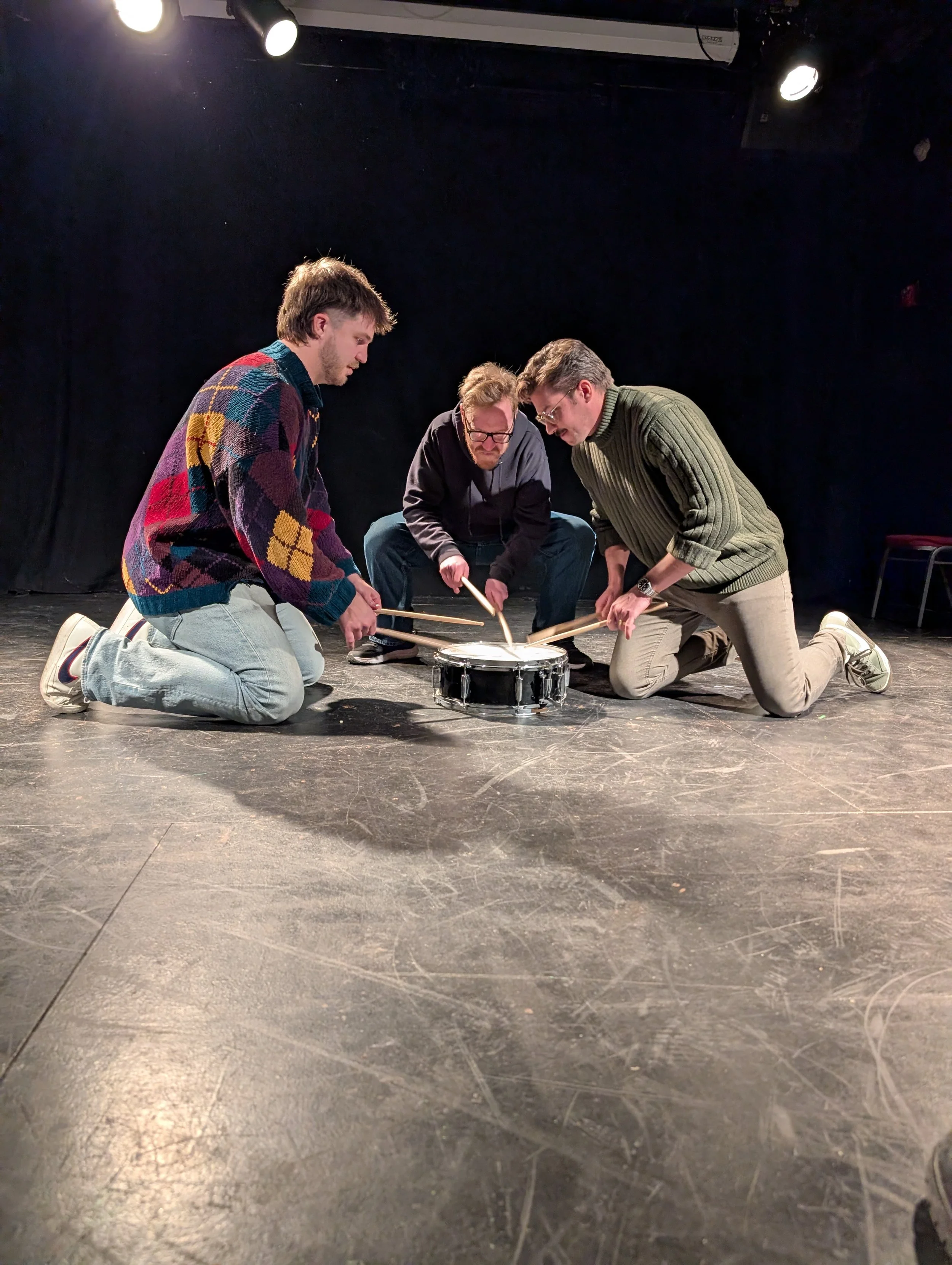 Three men kneeling on the stage floor, holding drumsticks and surrounding a small drum, appearing to prepare for a performance or rehearsal.