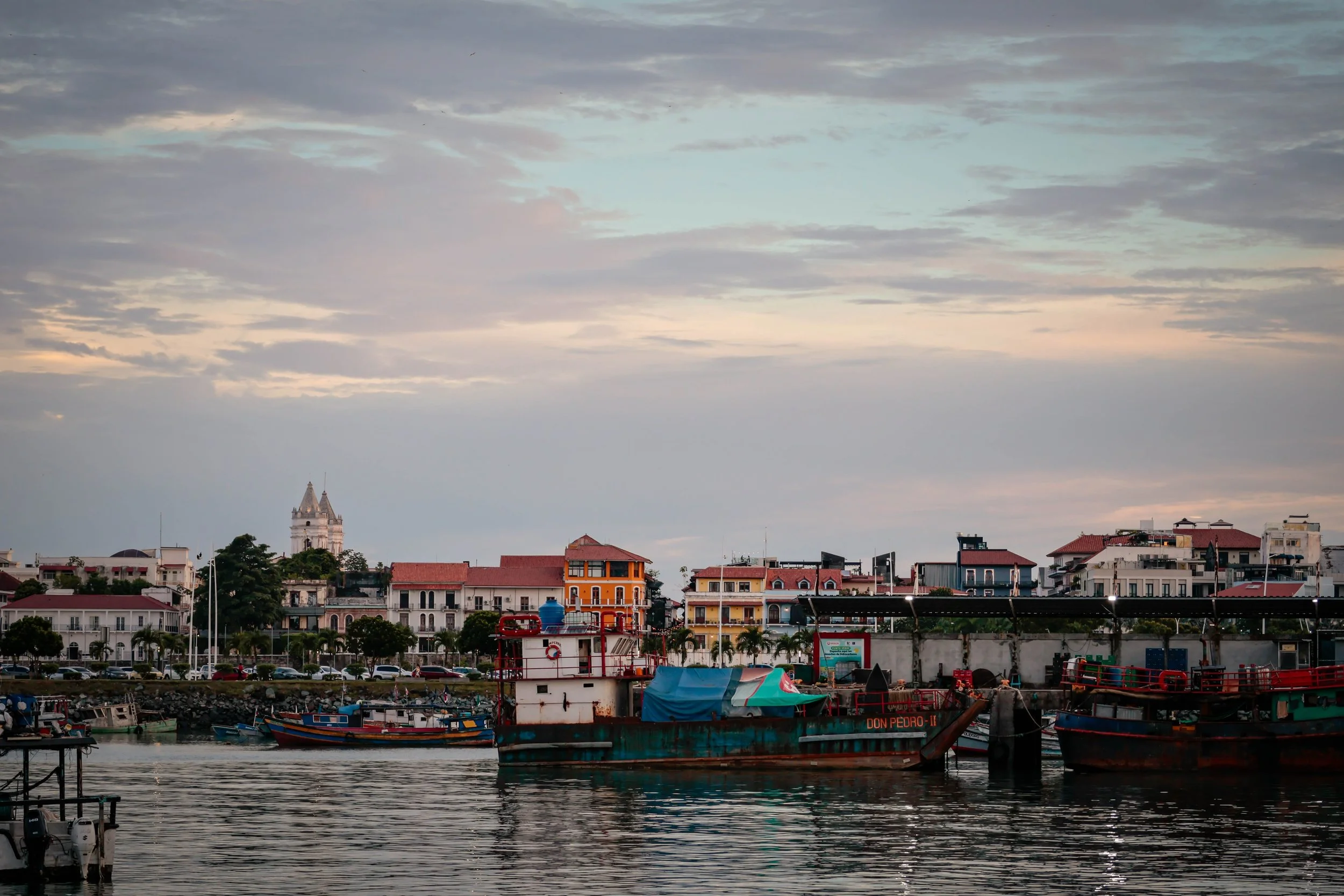 A harbor with boats docked in the water, colorful buildings and houses along the shoreline, and a cloudy sky overhead.