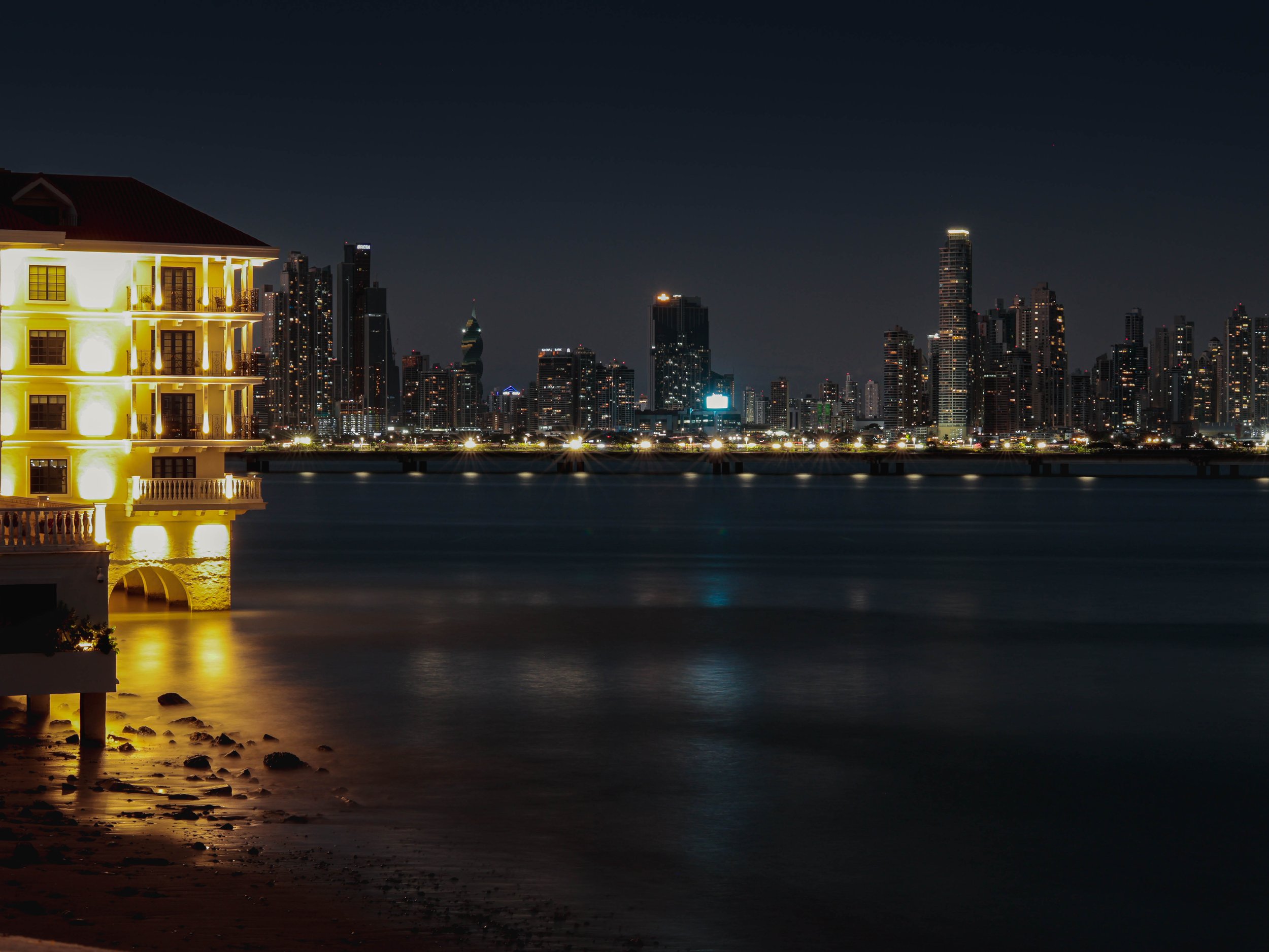Nighttime city skyline with illuminated buildings across a body of water, with a yellow-lit building in the foreground and rocks along the shore.