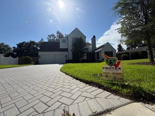 A house with a separate garage, a paved driveway, green lawn, and trees, with a PROTEX security sign in the front yard.