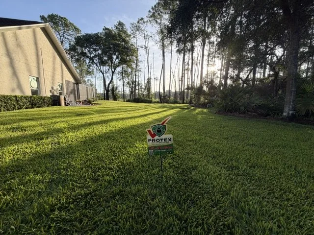 A well-maintained lawn with a small sign in the center that says "Protex" and has a logo with a green shield and red accents. In the background, there are trees and a building with a sloped roof, under a clear sky.