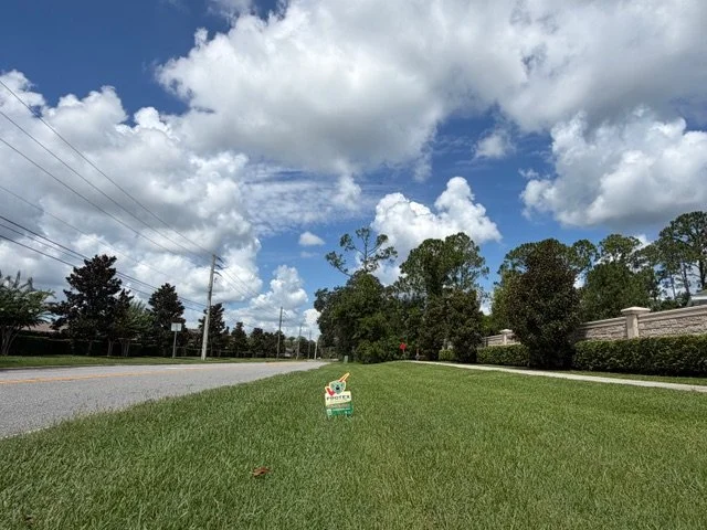 A grassy roadside with a small sign placed on the grass, under a partly cloudy sky, with trees and power lines in the background.