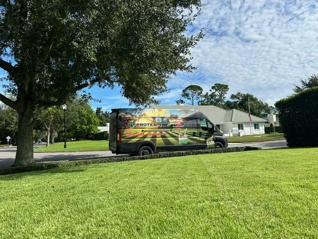 A black delivery truck with promotional graphics parked on a street in a suburban neighborhood, with a well-maintained lawn and large trees nearby.