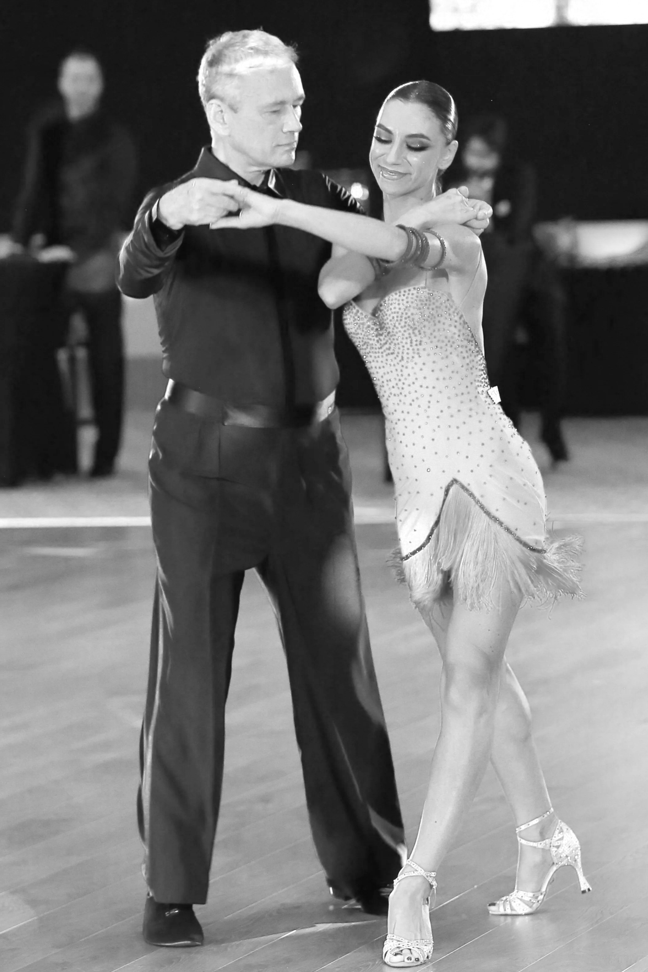 A man and woman dance together in a ballroom, with the man in dark clothing and the woman wearing a Fringed dress and high heels.