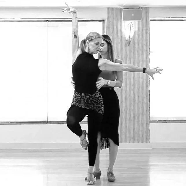 Two women dancing in a studio, one with an arm raised and leg bent, the other standing behind with arms around her waist.