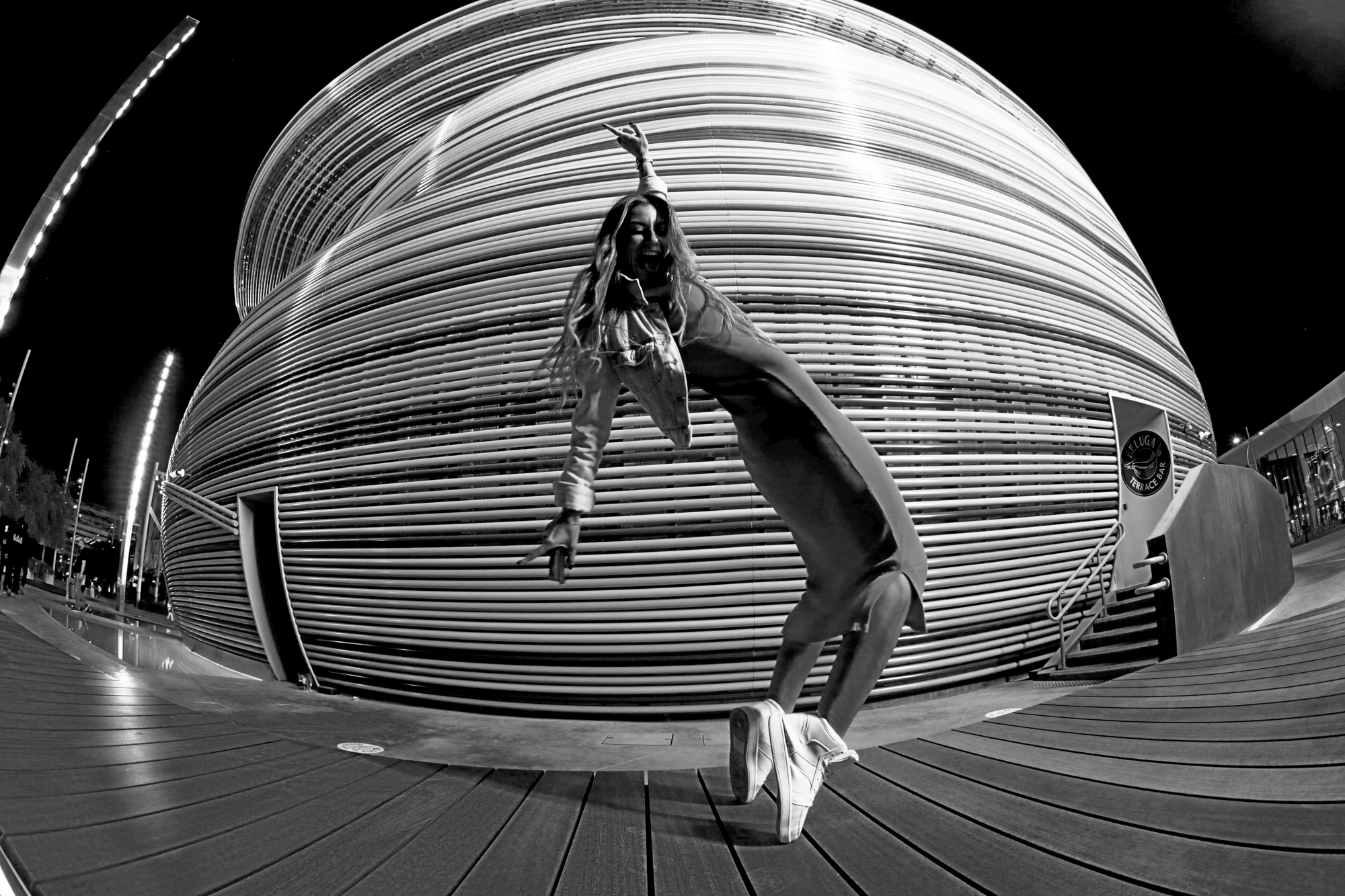 A woman dancing outdoors at night in front of a modern, curved, slatted building. She is in a dance pose with one arm raised and her mouth open, wearing high-heeled shoes, jeans, and a jacket.