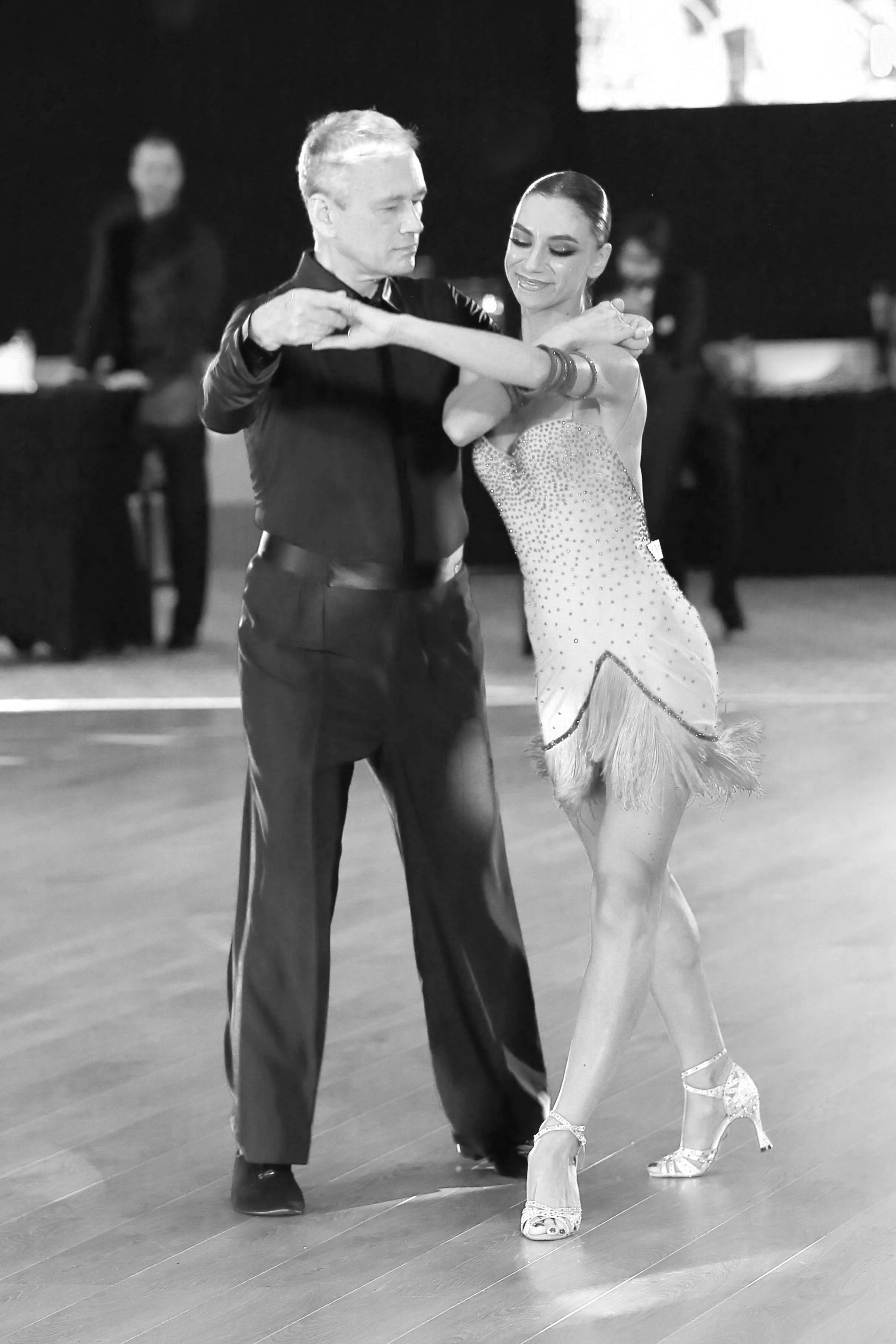 A couple performing a ballroom dance, the man is wearing a dark suit and the woman is in a sparkly dress with fringes and high heeled dance shoes, on a wooden floor with a dark background.